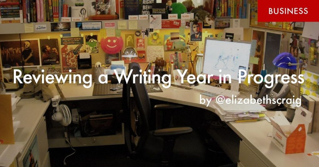 A desk with cube walls completely covered with colorful papers and posters is in the background and the foreground has the post title, "Reviewing the Writing Year in Progress" superimposed on it. 